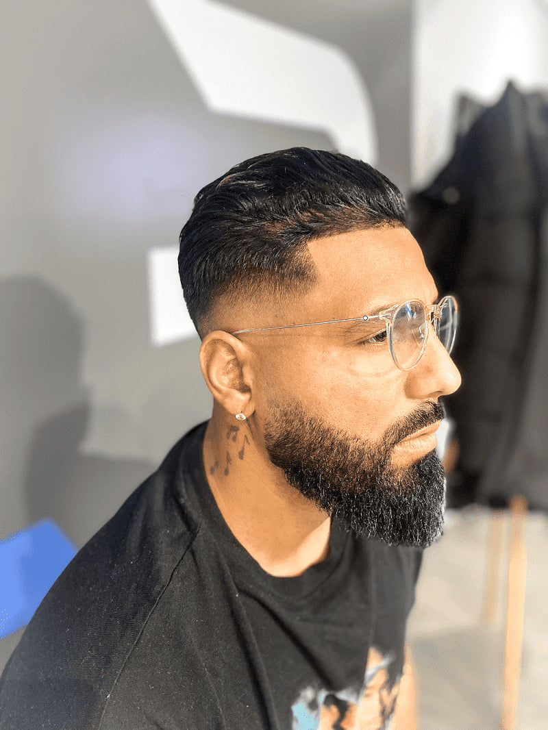 homme métis barbu et venant d'avoir un soin barbe portant des lunettes de vue transparentes, une boucle d'oreille et un tatouage dans le cou